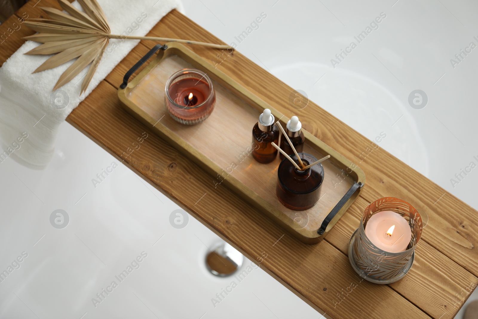 Photo of Wooden tray with cosmetic products, burning candles, and reed air freshener on bath tub in bathroom, above view