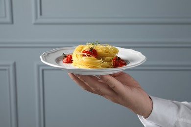 Photo of Waiter holding plate of tasty capellini with tomatoes and cheese near grey wall, closeup. Exquisite presentation of pasta dish