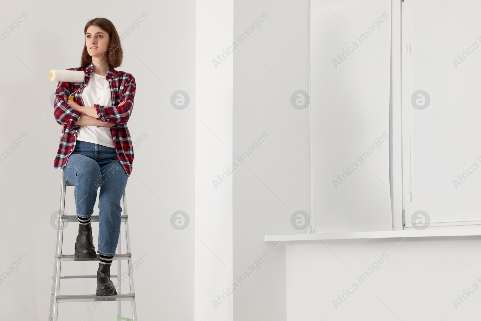 Photo of Young beautiful woman with roller on metal stepladder indoors. Room renovation