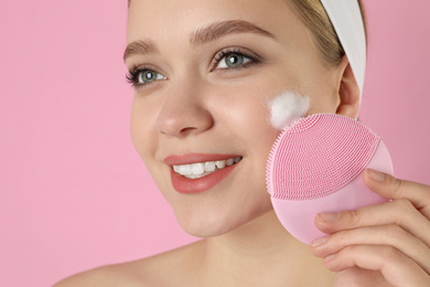 Photo of Young woman washing face with brush and cleansing foam on pink background. Cosmetic products