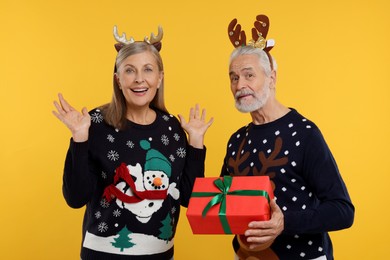 Couple in Christmas sweaters. Senior man presenting gift to his woman on orange background