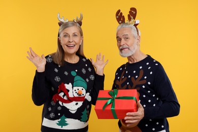 Photo of Couple in Christmas sweaters. Senior man presenting gift to his woman on orange background