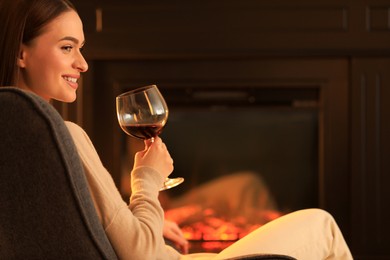 Photo of Young woman with glass of wine near fireplace indoors. Cozy atmosphere