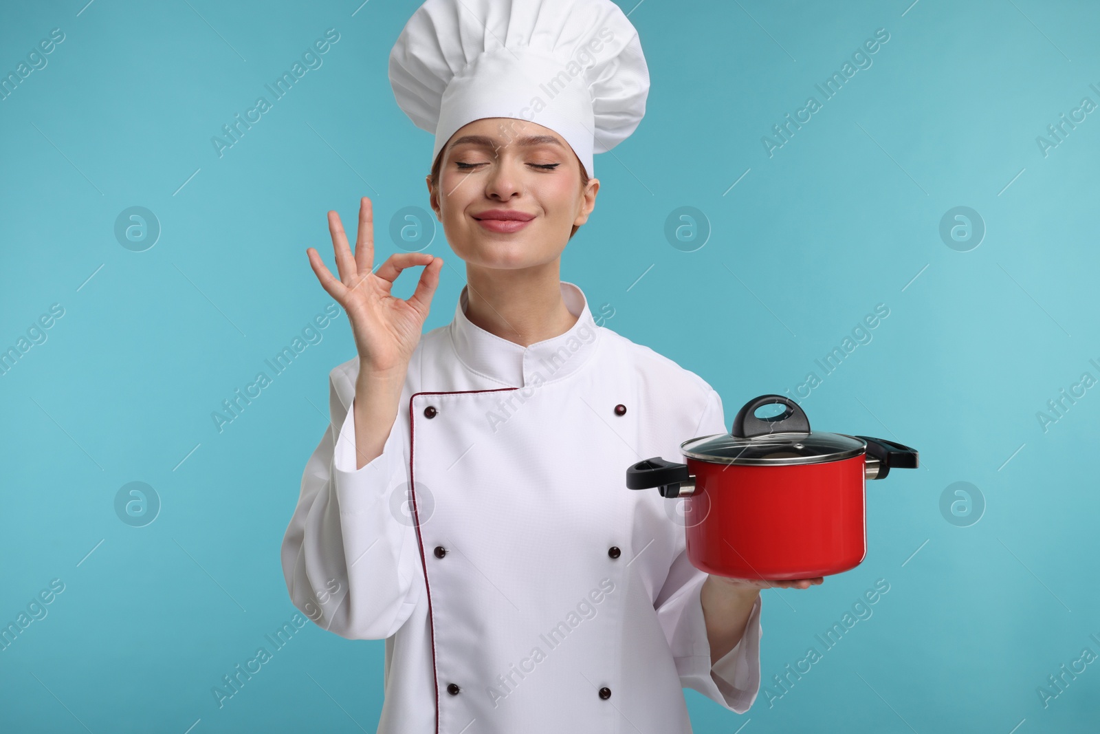 Photo of Happy woman chef in uniform holding cooking pot and showing perfect sign on light blue background
