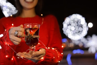 Woman with glass cup of mulled wine and garland at winter fair, closeup. Space for text