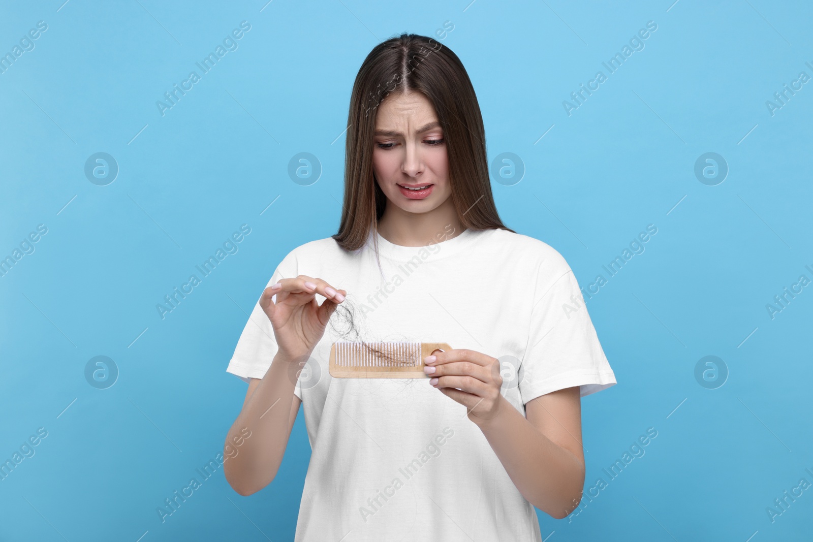 Photo of Emotional woman untangling her lost hair from comb on light blue background. Alopecia problem