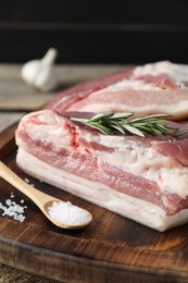 Photo of Pieces of raw pork belly, salt and rosemary on wooden board, closeup