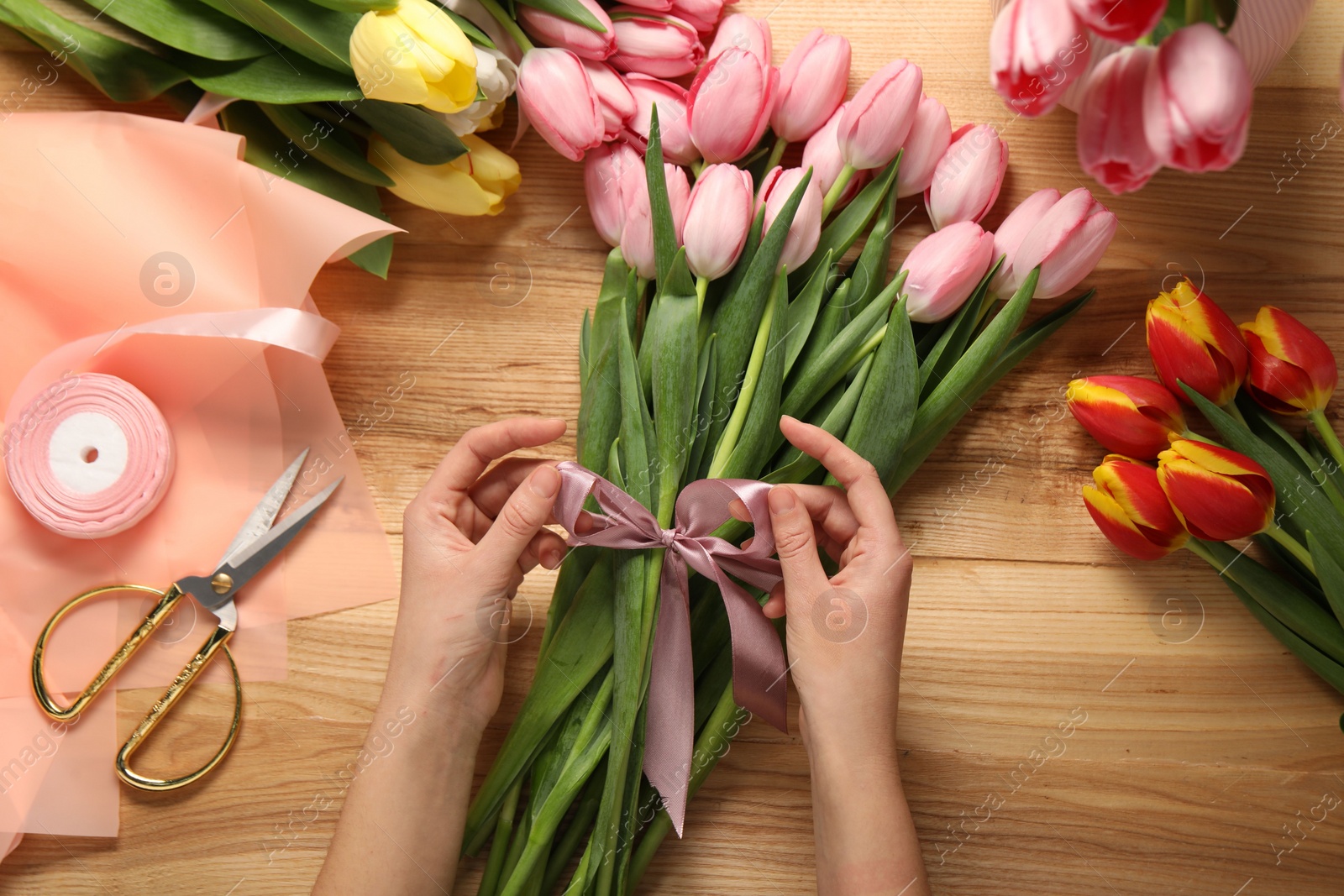 Photo of Woman making beautiful bouquet of fresh tulips and ribbon at wooden table, top view