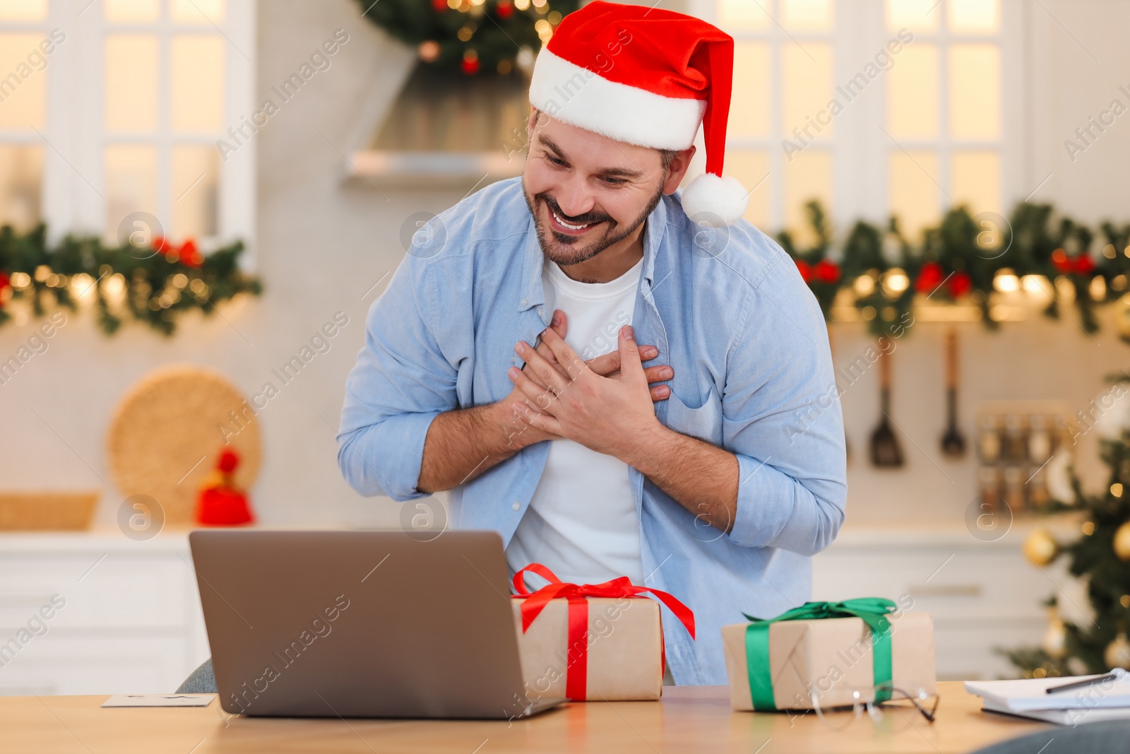 Photo of Celebrating Christmas online with exchanged by mail presents. Happy man in Santa hat thanking for gifts during video call on laptop at home