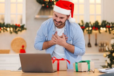 Celebrating Christmas online with exchanged by mail presents. Happy man in Santa hat thanking for gifts during video call on laptop at home