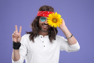 Photo of Stylish hippie man covering eye with sunflower and showing V-sign on violet background