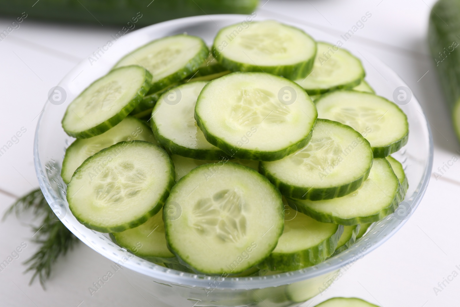 Photo of Cut cucumber in glass bowl on white table, closeup