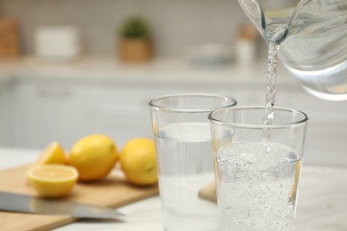 Pouring water from jug into glass on white table in kitchen, closeup