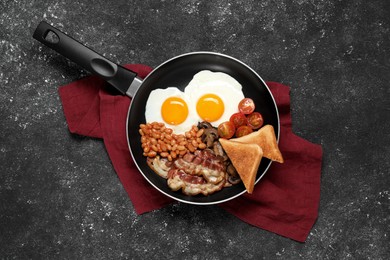 Frying pan with cooked traditional English breakfast on black textured table, top view