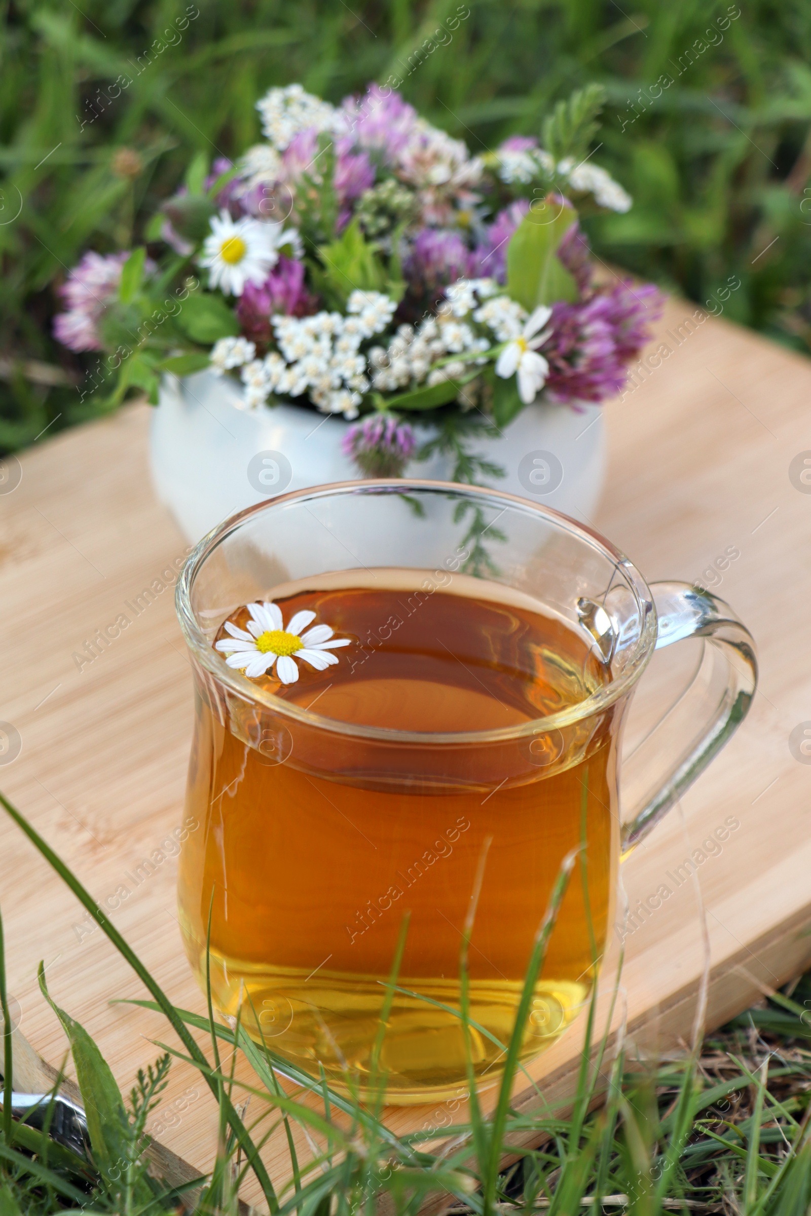 Photo of Cup of aromatic herbal tea and ceramic mortar with different wildflowers on green grass outdoors