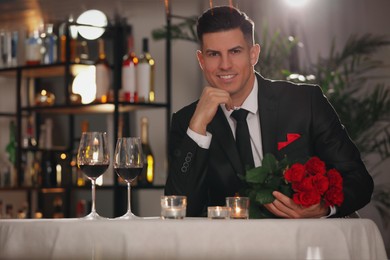 Photo of Happy man with roses waiting for his girlfriend in restaurant on Valentine's day