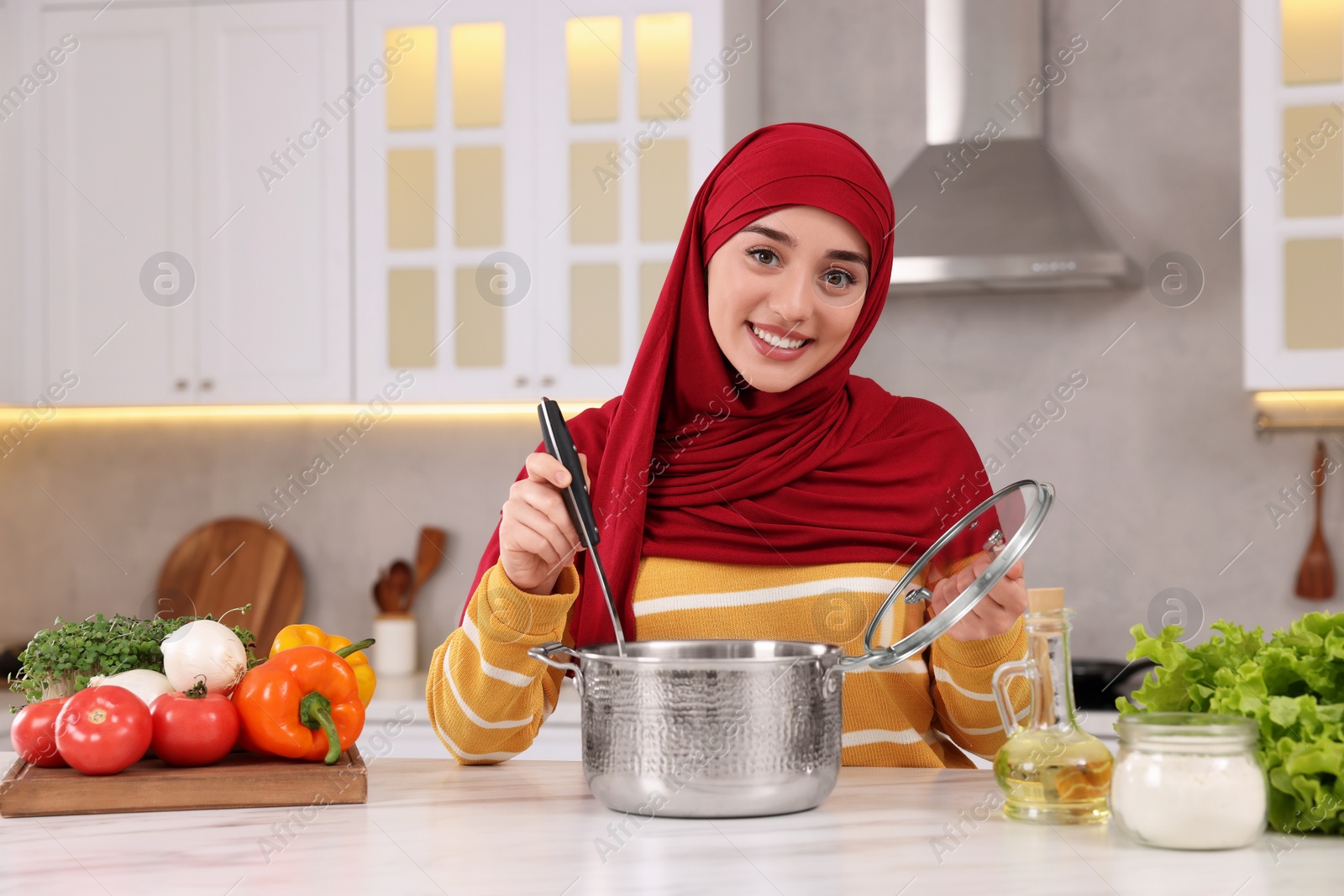 Photo of Muslim woman making delicious soup with vegetables at white table in kitchen