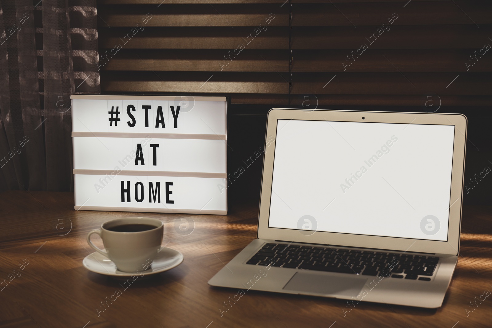 Photo of Laptop, cup of coffee and lightbox with hashtag STAY AT HOME on wooden table indoors. Message to promote self-isolation during COVID‑19 pandemic