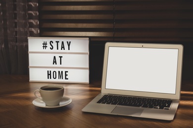 Photo of Laptop, cup of coffee and lightbox with hashtag STAY AT HOME on wooden table indoors. Message to promote self-isolation during COVID‑19 pandemic