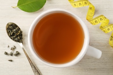 Photo of Cup of herbal diet tea and measuring tape on light wooden table, flat lay. Weight loss concept