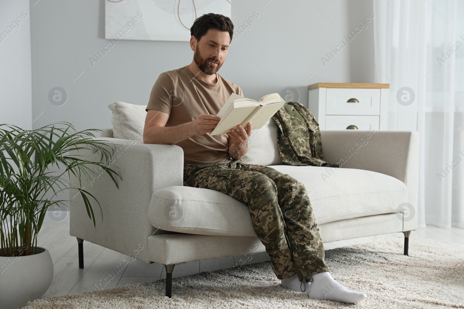 Photo of Soldier reading book on soft sofa in living room. Military service