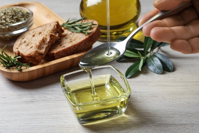 Photo of Woman pouring olive oil into spoon over bowl at wooden table, closeup