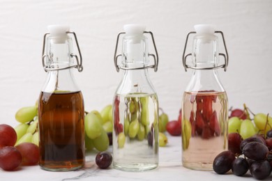 Photo of Different types of vinegar in bottles and grapes on light marble table, closeup