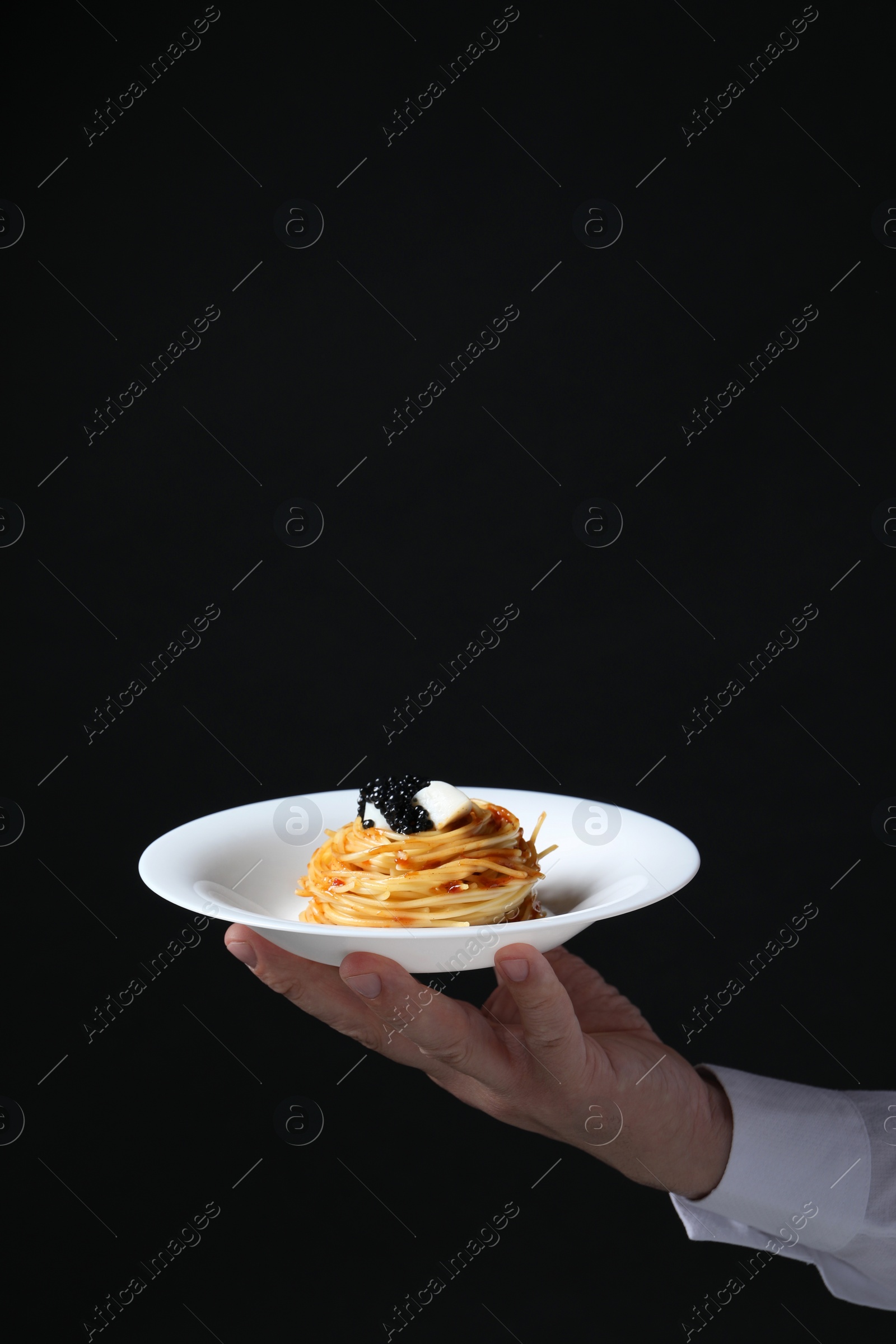 Photo of Waiter holding plate of tasty spaghetti with tomato sauce and black caviar on dark background, closeup and space for text. Exquisite presentation of pasta dish