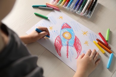 Photo of Little boy drawing rocket with soft pastel at wooden table, closeup. Child`s art