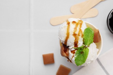 Photo of Scoops of tasty ice cream with caramel sauce, mint and candies on white tiled table, top view. Space for text