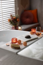 Photo of White wooden tray with glass of rose wine, book and burning candles on bathtub in bathroom