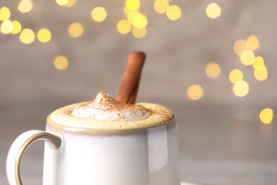 Photo of Cup of delicious eggnog with cinnamon on blurred background against festive lights, closeup