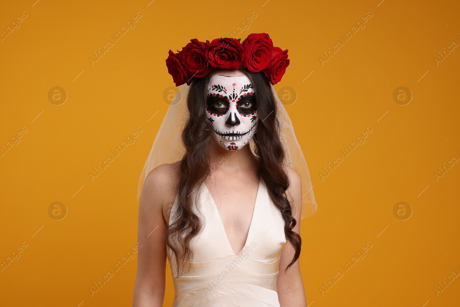 Photo of Young woman in scary bride costume with sugar skull makeup and flower crown on orange background. Halloween celebration