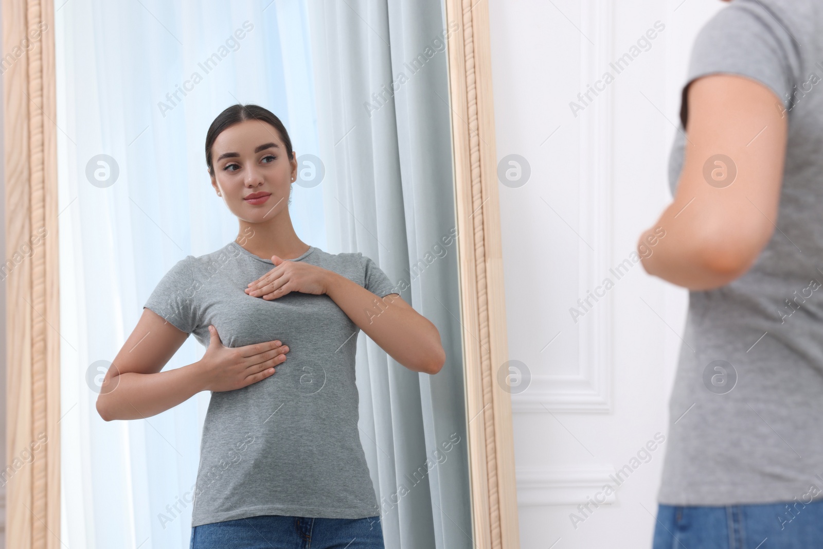 Photo of Beautiful young woman doing breast self-examination near mirror indoors