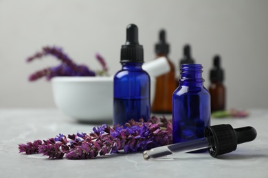 Photo of Bottles of sage essential oil and flowers on grey table