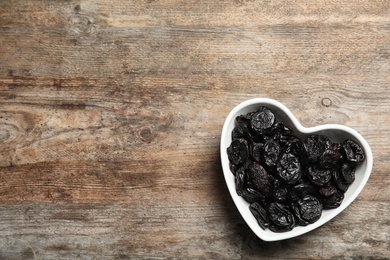 Photo of Heart shaped bowl of sweet dried plums on wooden background, top view with space for text. Healthy fruit