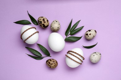 Beautifully decorated Easter eggs and green leaves on lilac background, flat lay