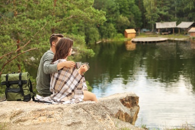 Photo of Cute couple with mugs and plaid near lake. Camping season