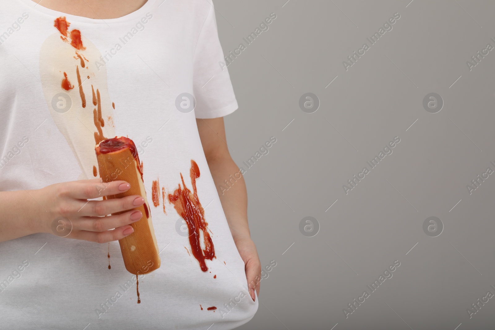 Photo of Woman holding hotdog and showing stain from sauce on her shirt against light grey background, closeup. Space for text