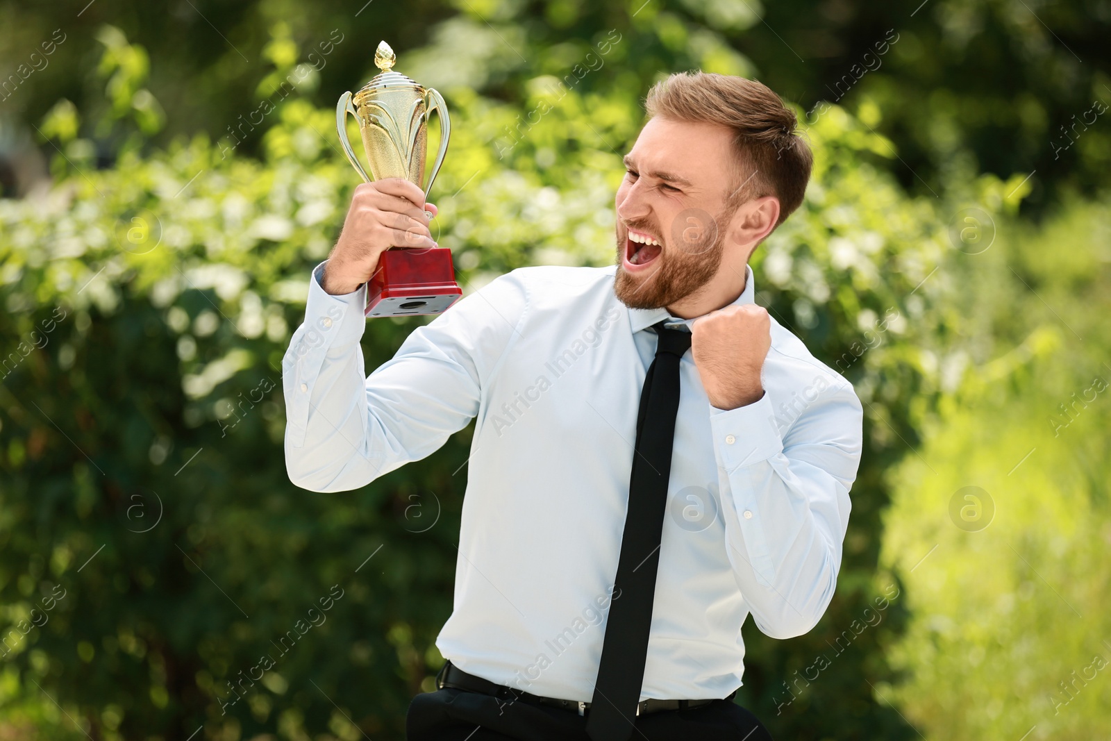 Photo of Portrait of happy young businessman with gold trophy cup in green park