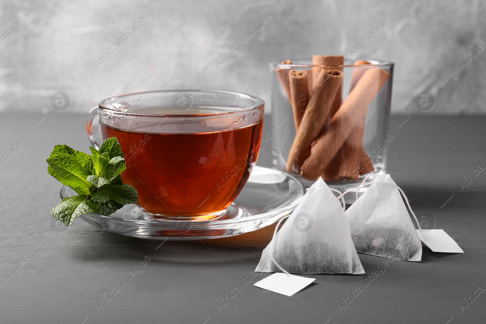 Photo of Tea bags, cup of hot drink, mint and cinnamon sticks on grey wooden table