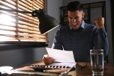 Excited man reading letter at wooden table in dark room