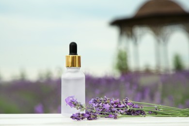 Photo of Bottle of essential oil and lavender flowers on white wooden table in field, space for text