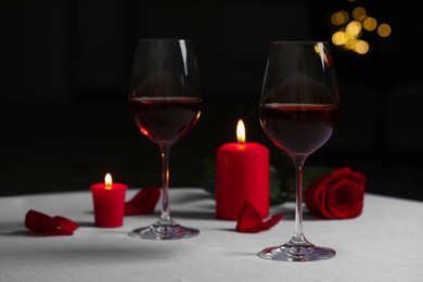 Photo of Glasses of red wine, rose flower and burning candles on white table against blurred lights. Romantic atmosphere