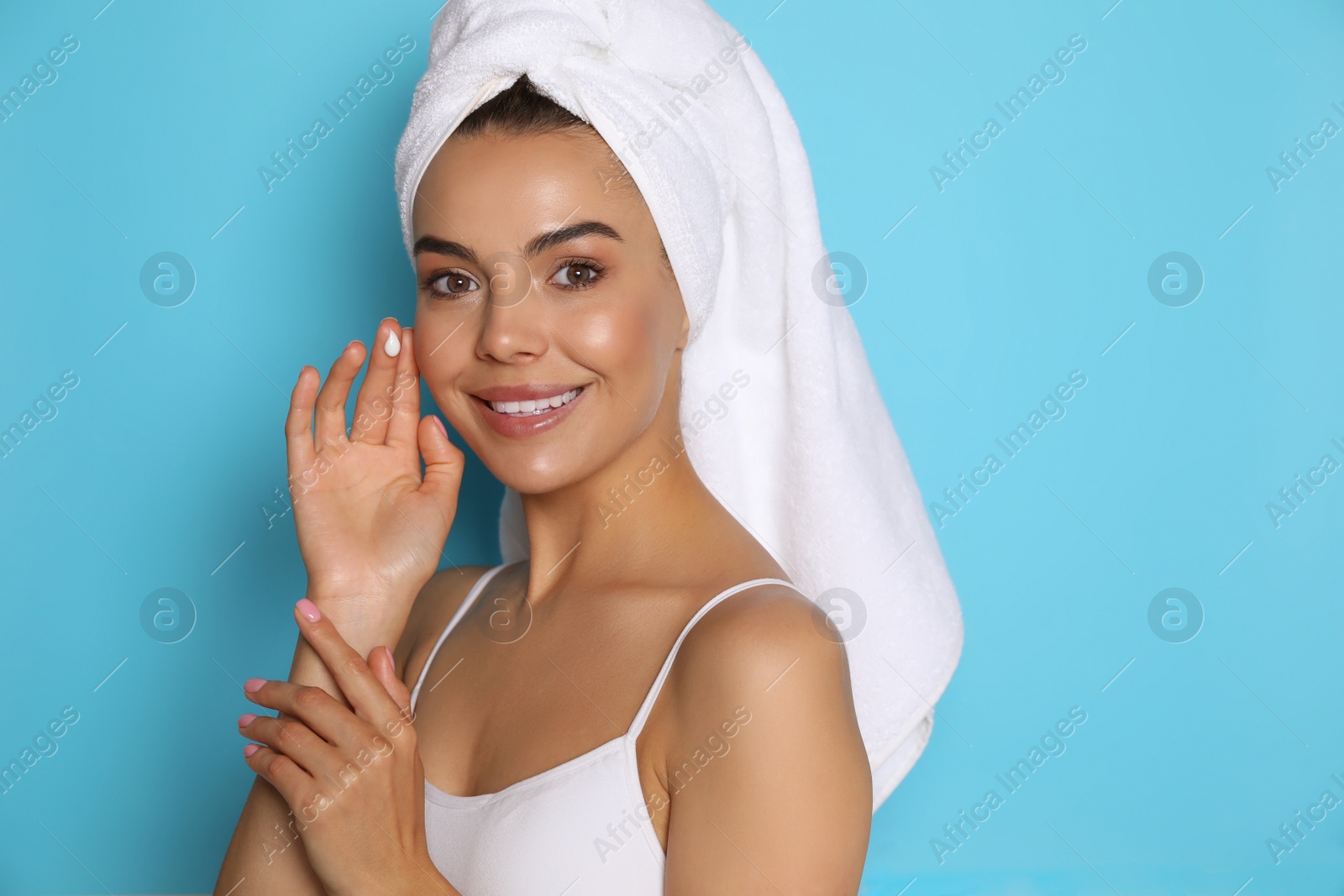 Photo of Beautiful young woman with towel applying cream on face against light blue background