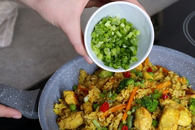 Photo of Woman adding cut green onion to rice with meat and vegetables in frying pan, closeup