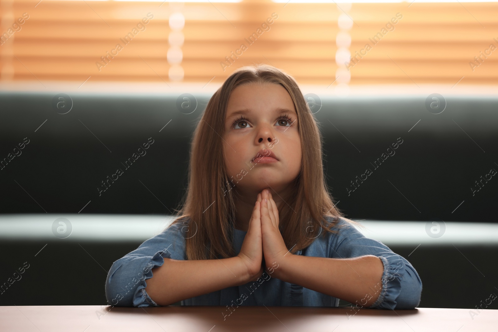 Photo of Cute little girl with hands clasped together praying at table indoors