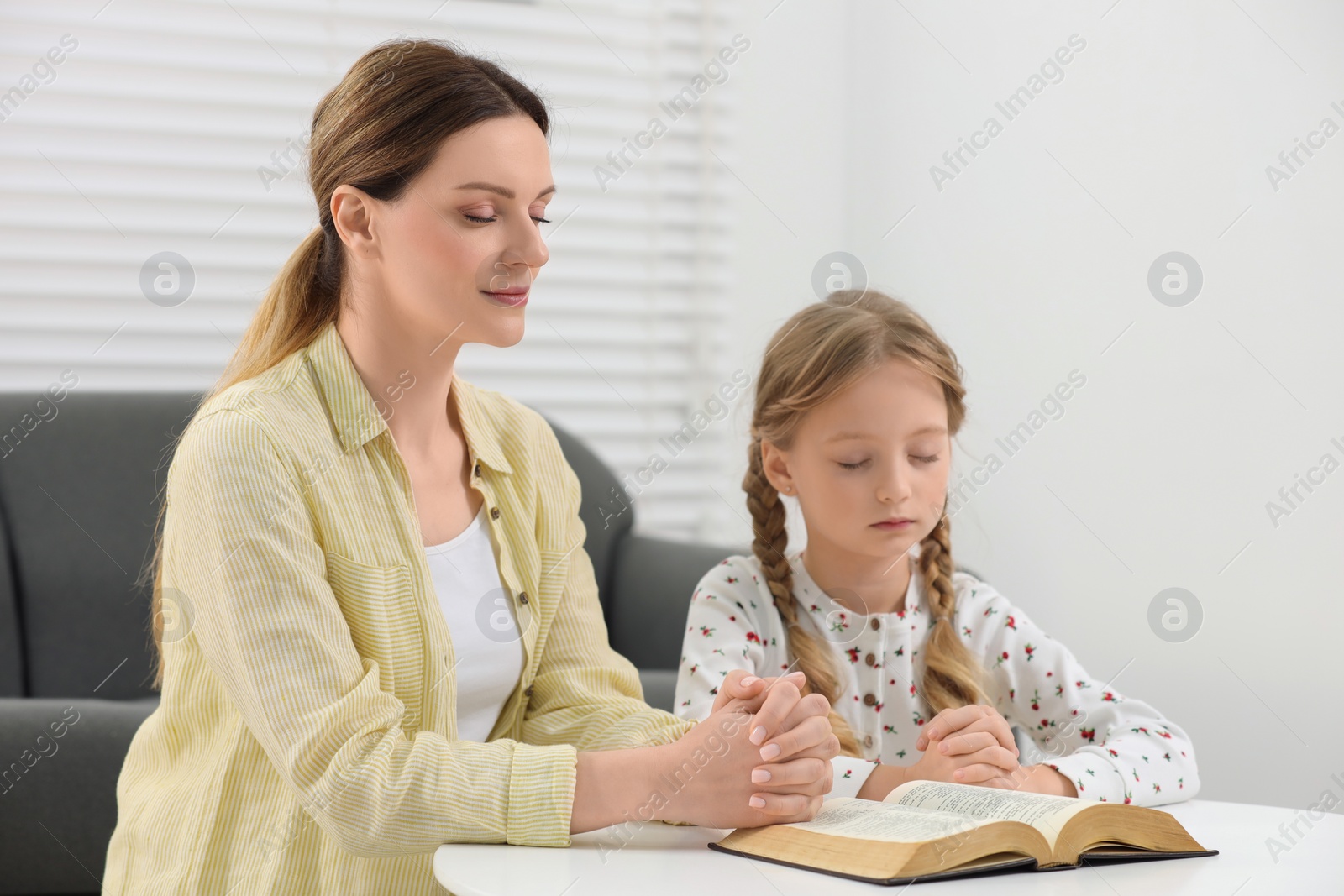 Photo of Girl and her godparent praying over Bible together at table indoors