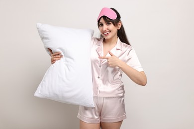Happy woman in pyjama and sleep mask holding pillow on light grey background