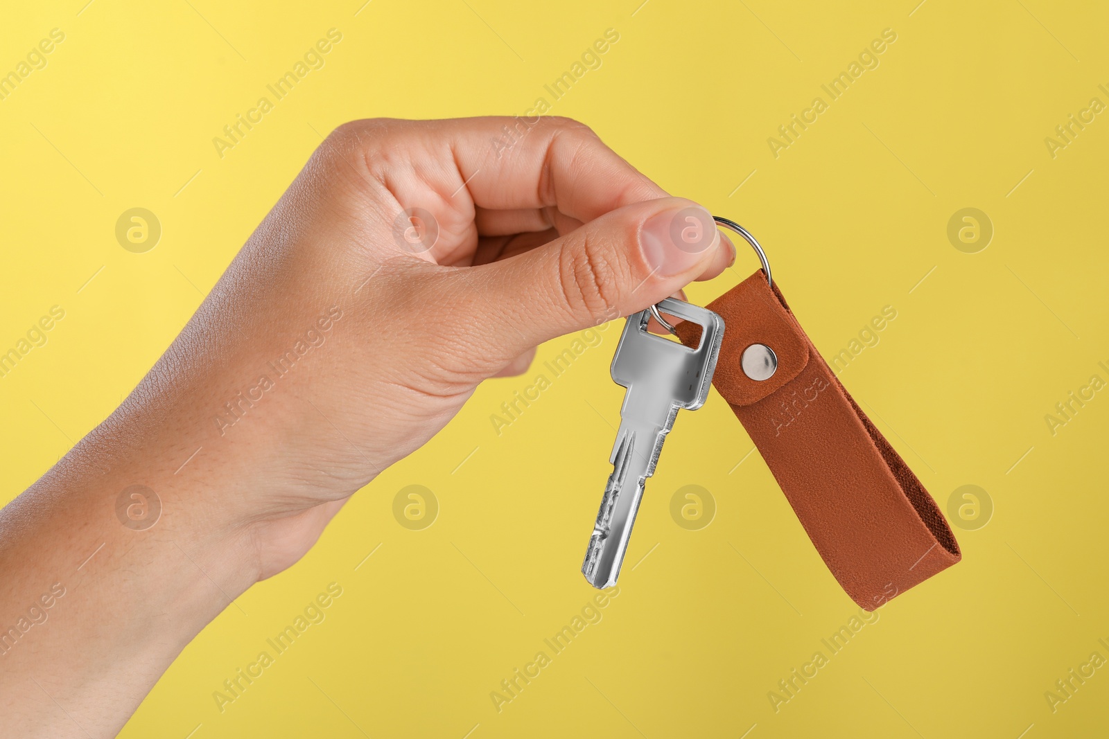 Photo of Woman holding key with leather keychain on yellow background, closeup